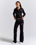 A woman modeling a black athletic tracksuit with white contrast stitching and white logo text on the chest and thigh, photographed against a light gray studio background.