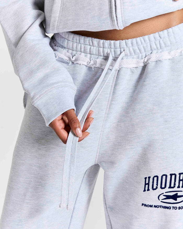 Close-up of a person wearing light heather gray sweatpants with a drawstring waist and a navy graphic print on the left leg that reads 'HOODRICH'