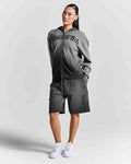 A woman modeling a dark gray two-piece tracksuit featuring a zip-up hoodie with 'Hoodrich' printed across the chest and matching shorts with 'AlexRich' printed down the leg, paired with white sneakers against a light gray studio background.