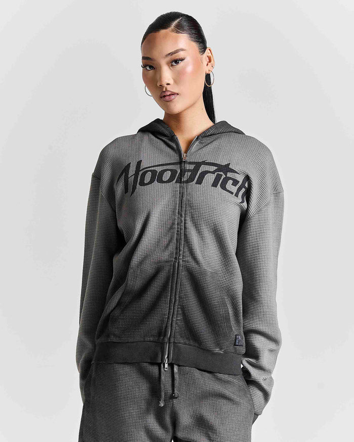 A woman modeling a dark gray waffle-knit zip-up hoodie with a black 'Hoodrich' logo across the chest, paired with matching gray pants, against a light gray studio background.