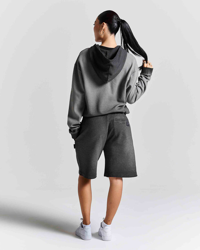 A woman shown from the back wearing a gray and black hooded sweatshirt, dark gray shorts, and white sneakers against a light gray studio background.