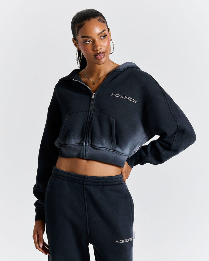 Model wearing a black cropped zip-up hoodie with a Hoodrich logo on the chest.