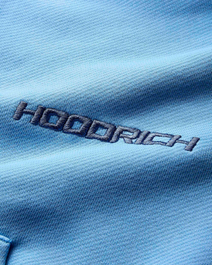 Close-up of the dark blue Hoodrich logo embroidered on the blue fabric of the hoodie.