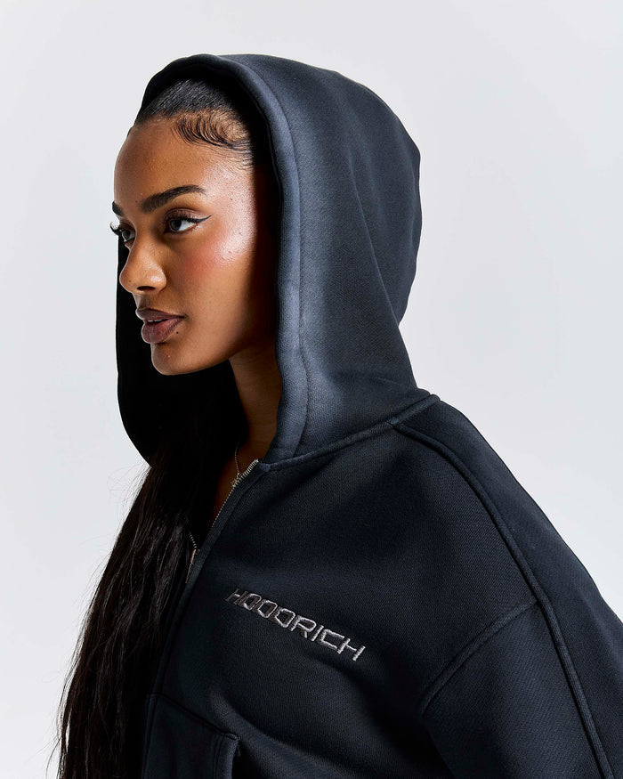 A woman wearing a black hooded sweatshirt with a silver 'HOODRICH' logo on the chest, shown in a side profile against a light gray background.