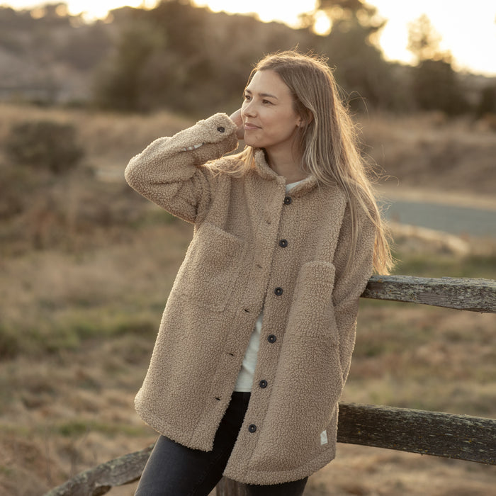 A woman in the Pladra Women's Headlands Sherpa Shacket in Almond Tan smiles warmly while leaning on a wooden fence outdoors.