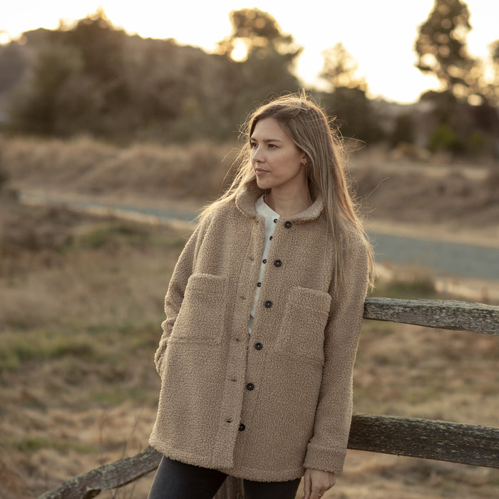 Woman in Pladra Women's Headlands Sherpa Shacket - Almond Tan stands by a wooden fence at sunset in a rural setting.