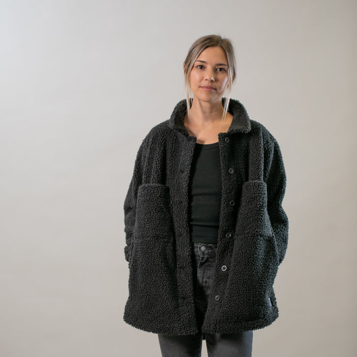 A person stands against a plain light background, wearing an oversized Women's Headlands Sherpa Shacket in Onyx Black with plaid flannel lining, layered over a black top and gray pants. Their hair is pulled back and they have a neutral expression.