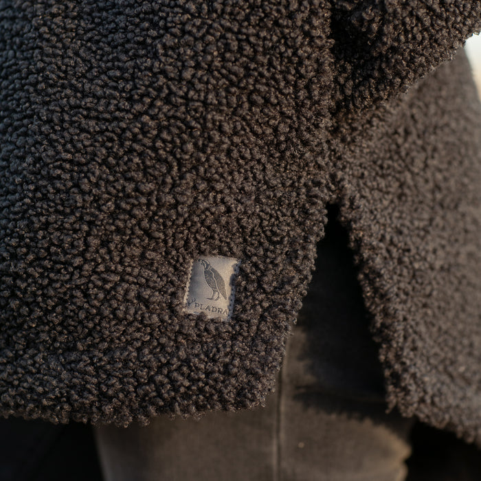 Close-up of a textured, dark Women's Headlands Sherpa Shacket in Onyx Black, featuring a plaid flannel lining and a small fabric label with a bird and the Plandra brand name stitched near the hem.