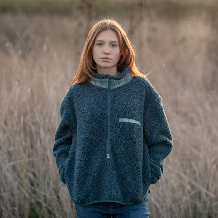 A young woman with long red hair stands in a field of tall grass, wearing the Women's Jenner Sherpa Fleece- Atlantic Blue with recycled polyester, hands in her pockets, and looks at the camera with a neutral expression.