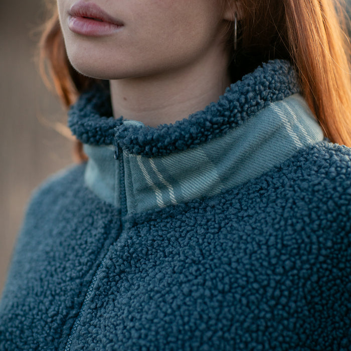 Close-up of a woman with long red hair wearing the Women's Jenner Sherpa Fleece in Atlantic Blue, made from recycled polyester with a high collar, visible zipper, and cozy plaid flannel lining; only her lower face and neck are shown.