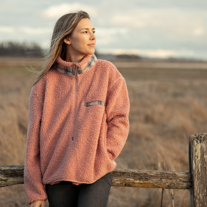 A woman in the Women's Jenner Sherpa Fleece - Coral Pink stands by a wooden fence in an open field, gazing calmly into the distance under a cloudy sky.