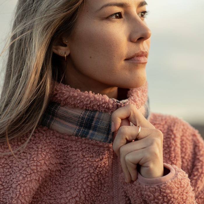 A woman in a coral pink Women's Jenner Sherpa Fleece with plaid flannel lining stands outdoors, gazing thoughtfully into the distance. She holds the collar with one hand, her light brown hair loose and hoop earrings visible.