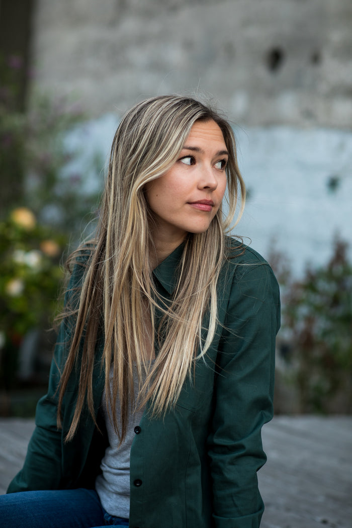 A young woman sits outdoors in a Pladra Women's Leon Workhorse Shirt - Fir Green Pincord, looking to her left.