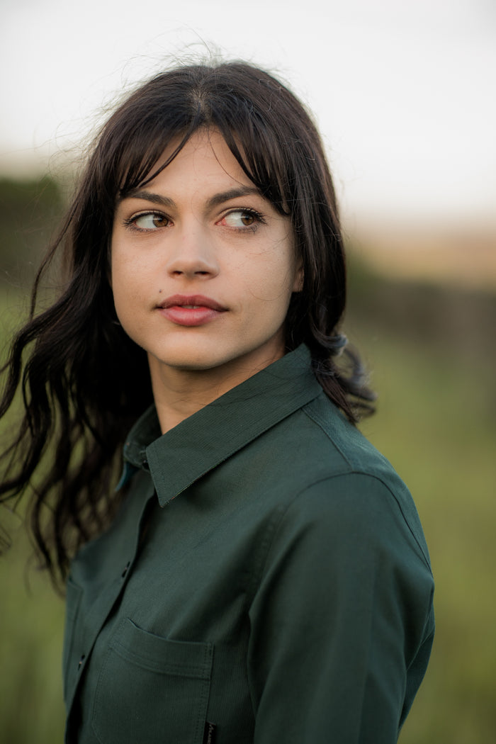 Woman with dark hair in a Pladra Women's Leon Workhorse Shirt - Fir Green Pincord stands outdoors, looking left.