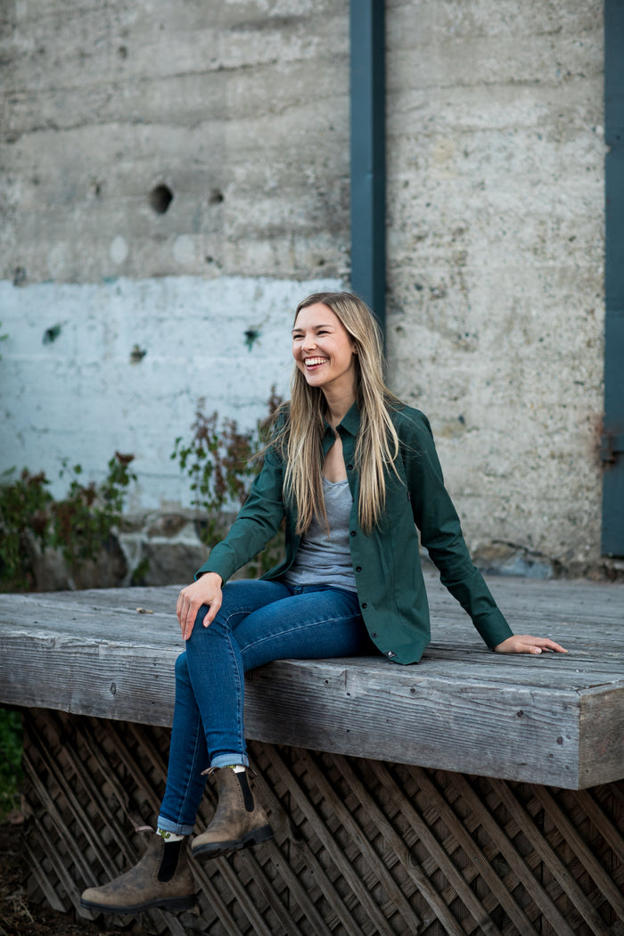 A woman on a bench wears Pladra's Women's Leon Workhorse Shirt in Fir Green Pincord, smiling by a concrete wall.