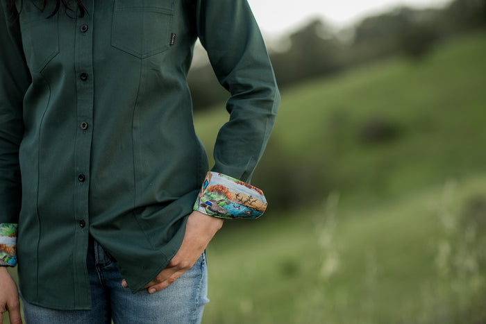 A person wearing Pladra’s Women's Leon Workhorse Shirt–Fir Green Pincord and jeans stands outdoors in a grassy field.