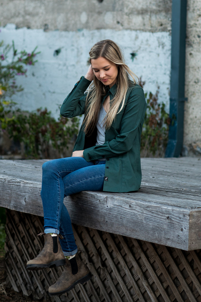 A woman in a Pladra Women's Leon Workhorse Shirt- Fir Green Pincord and jeans sits on a wooden platform, looking down.