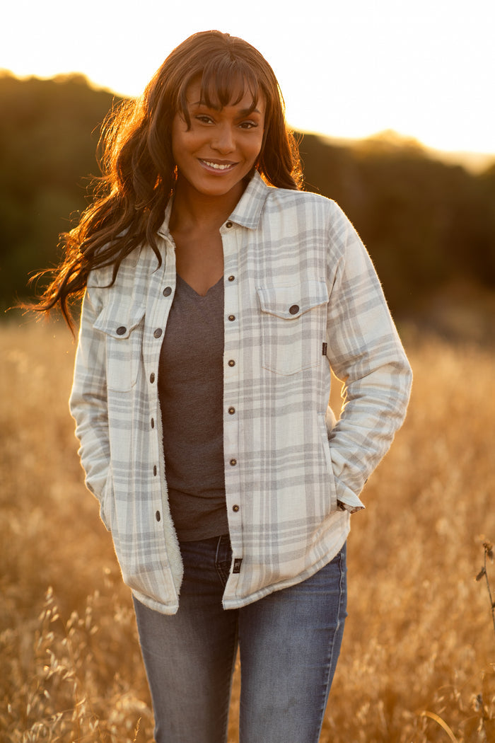 Woman smiling outdoors in a field, wearing the Pladra Women's Northwood Sherpa Insulated Flannel Jacket in Cloud Grey.