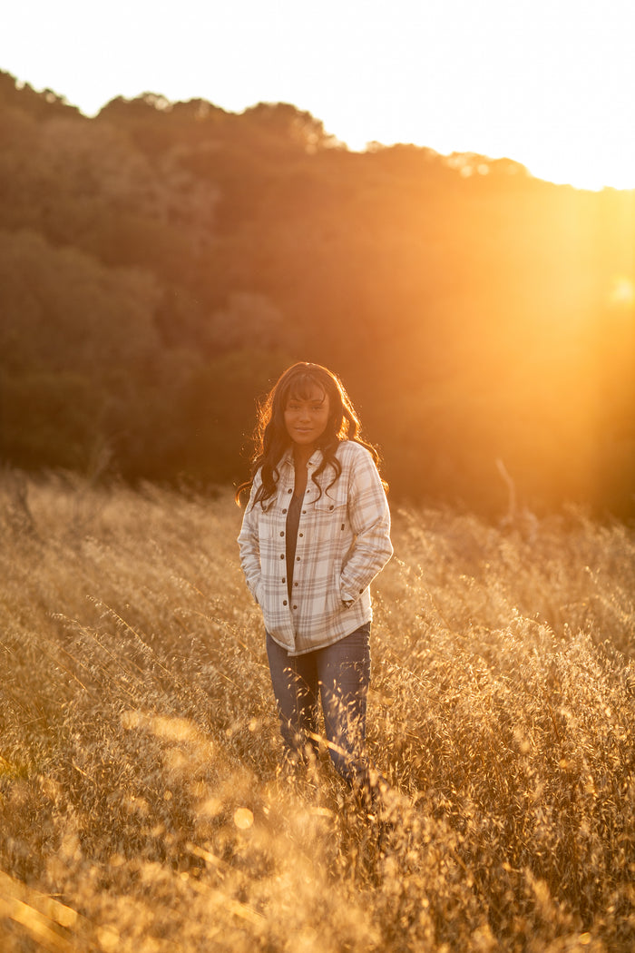 A woman stands in a sunlit field wearing the Pladra Women's Northwood Sherpa Insulated Flannel Jacket in Cloud Grey.