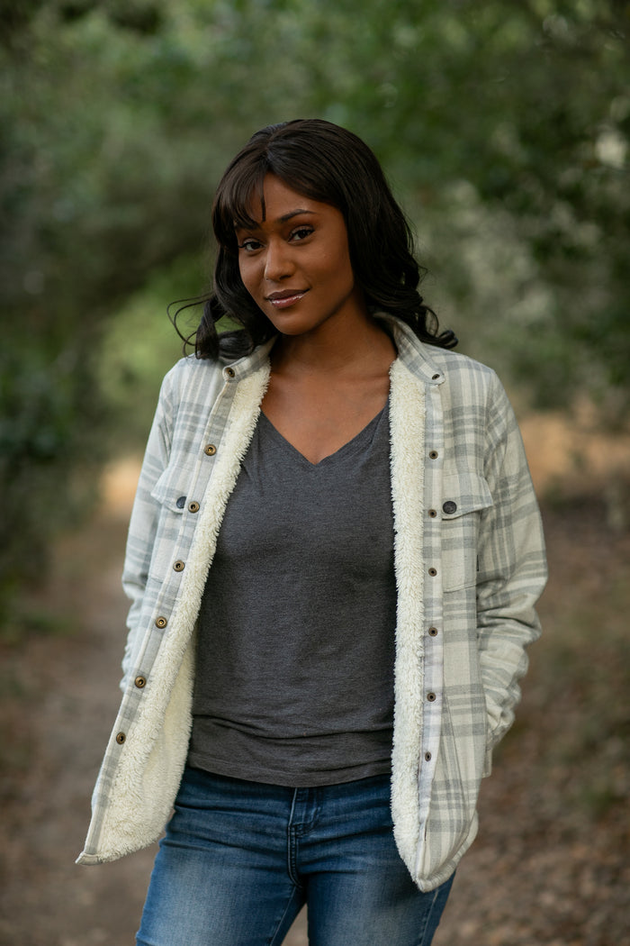A woman in Pladra's Women's Northwood Sherpa Insulated Flannel Jacket—Cloud Grey stands on a wooded path, smiling.