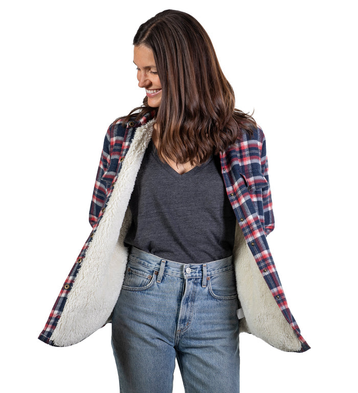 A woman smiles, looking down to her left, wearing blue jeans and the Women's Northwood Sherpa Insulated Flannel Jacket in Eagle Blue with a cozy white fleece lining, standing against a white background.