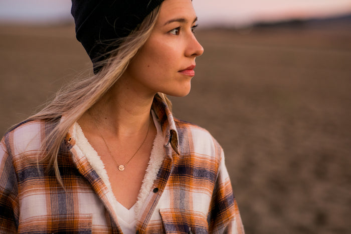Woman in a Pladra Women's Northwood Sherpa Flannel Jacket in Peak White and beanie gazes outside at sunset.
