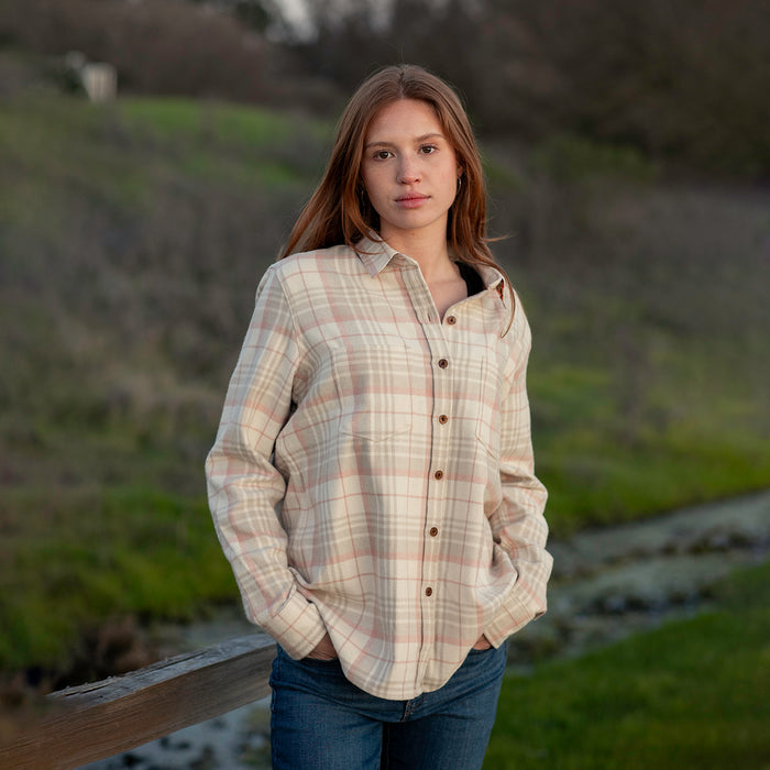 A young woman wears the Women's Relaxed Every Day Elite Flannel Shirt in Lighthouse White with jeans, standing outdoors by a wooden fence amid grass and trees, hands in her pockets.