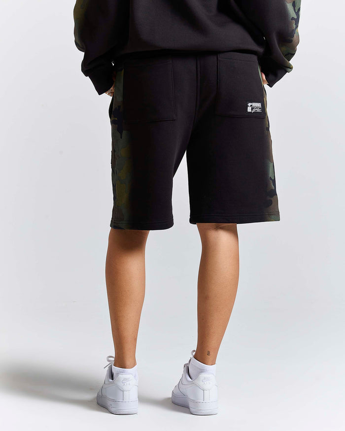 A person shown from the back wearing black shorts with camouflage side panels, a black sweatshirt, and white sneakers against a light gray studio background.
