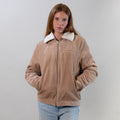 Woman with long hair wears a Pladra Women's Stinson Jacket–Oak Tan and blue jeans, standing against a plain background.