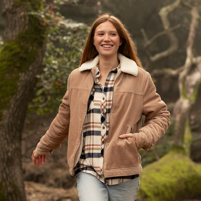 Woman in Pladra Women's Stinson Jacket - Oak Tan walks outdoors among mossy trees.