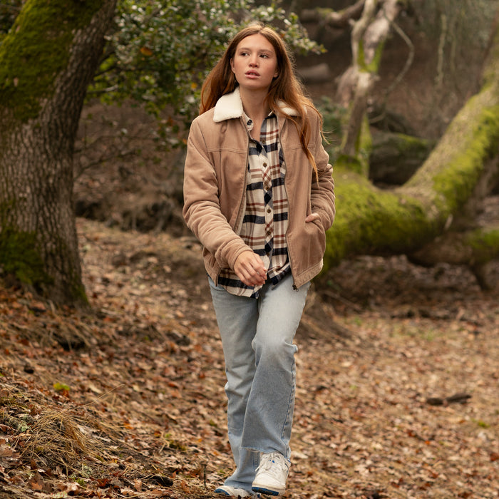 A woman in Pladra’s Women’s Stinson Jacket in Oak Tan walks through a forest with fallen leaves.
