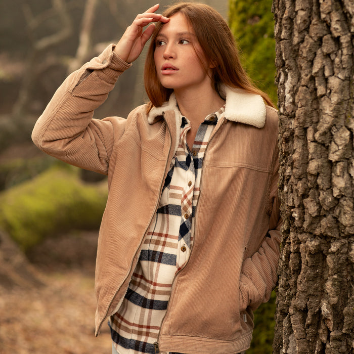 Woman in Pladra’s Women's Stinson Jacket in Oak Tan stands by a tree, gazing into the distance.
