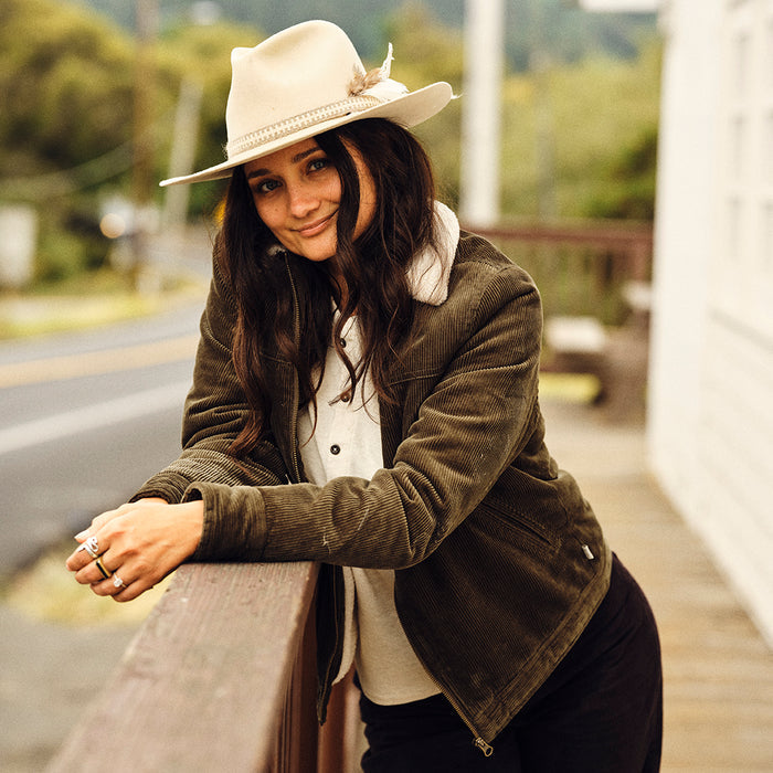A woman with long dark hair smiles outdoors, leaning on a wooden railing. She wears rings, a light wide-brimmed hat, and the Women's Stinson Jacket in Pine Green. A road and greenery are visible in the background.