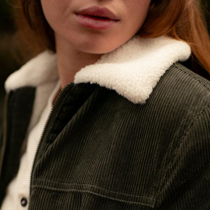 Close-up of a person wearing the Women's Stinson Jacket in Pine Green, featuring a white sherpa collar. Only the lower half of their face and upper chest are visible; background is blurred.
