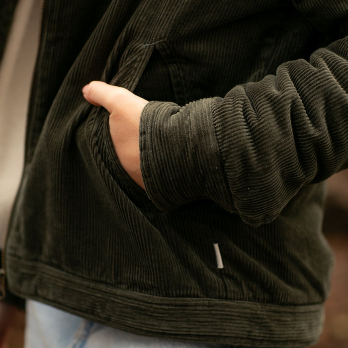 A person wears the Women's Stinson Jacket in Pine Green with a hand in the pocket. Only the torso and part of the arm are visible. Light-colored pants are also worn.