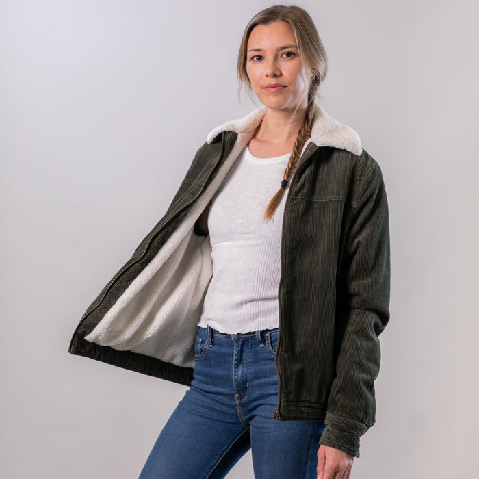 A woman with a braid wears the Women's Stinson Jacket—Pine Green, featuring white sherpa lining—over a white top and blue jeans. She stands against a plain light background, jacket open to reveal the interior lining.