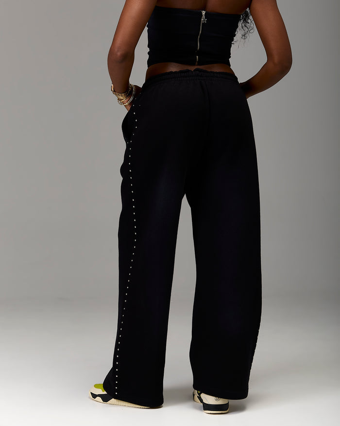 A model from the back wearing black wide-leg pants with a decorative line of silver studs down the side, paired with a black strapless top and sneakers, set against a plain gray studio background.