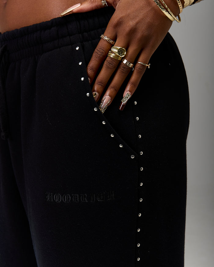 Close-up of a person wearing black jogger sweatpants with silver stud embellishments down the pocket and a subtle embossed logo on the thigh. The person's hand and decorated nails are resting on the waistband, with gold rings and bracelets visible against a neutral gray background.