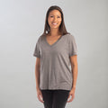 A woman with long brown hair smiles against a light gray background, wearing the Women's V-Neck Tri-Blend Tee in Charcoal Grey Heather, featuring an oversized silhouette and paired with black pants.
