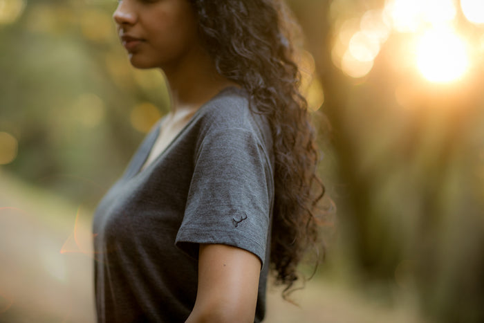A woman with long, curly hair stands outdoors in a Women's V-Neck Tri-Blend Tee—Charcoal Grey Heather. Sunlight illuminates the soft jersey and highlights her profile against a blurred backdrop of trees.