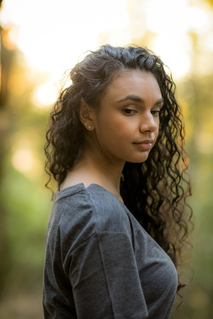 A young woman with long curly hair stands outdoors in soft sunlight, wearing a Women's V-Neck Tri-Blend Tee in Charcoal Grey Heather. She looks over her shoulder calmly, with a blurred background of warm, natural colors.
