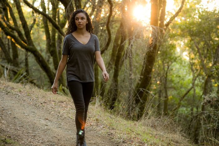 A woman with curly hair, wearing an oversized Women's V-Neck Tri-Blend Tee in Charcoal Grey Heather and pants, walks along a sunlit forest path surrounded by trees and foliage.