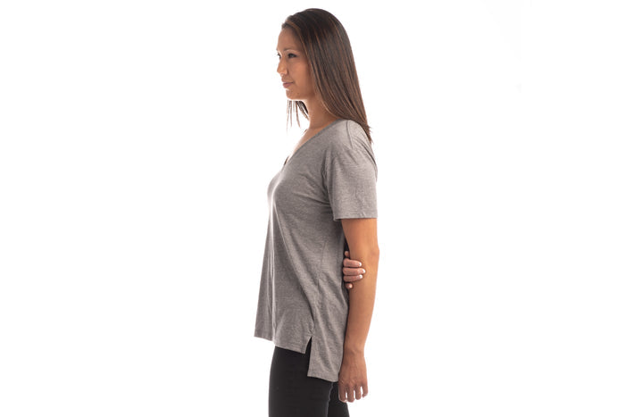 A woman with straight brown hair stands in profile against a white background, wearing a Women's V-Neck Tri-Blend Tee- Charcoal Grey Heather and black pants, looking forward with a neutral expression.
