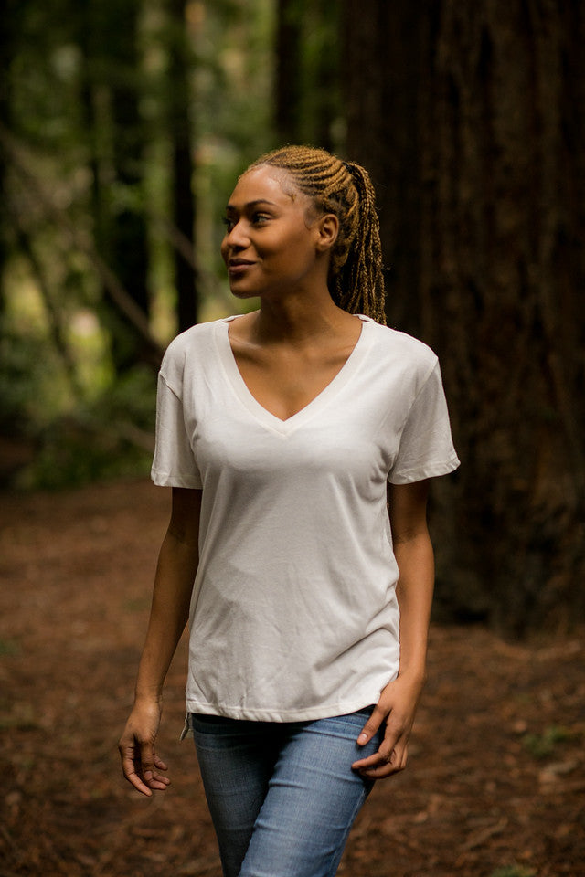 Woman in a Pladra Women's V-Neck Tri-Blend Tee - Marshmallow White Heather walks in a sunlit forest among tall trees.