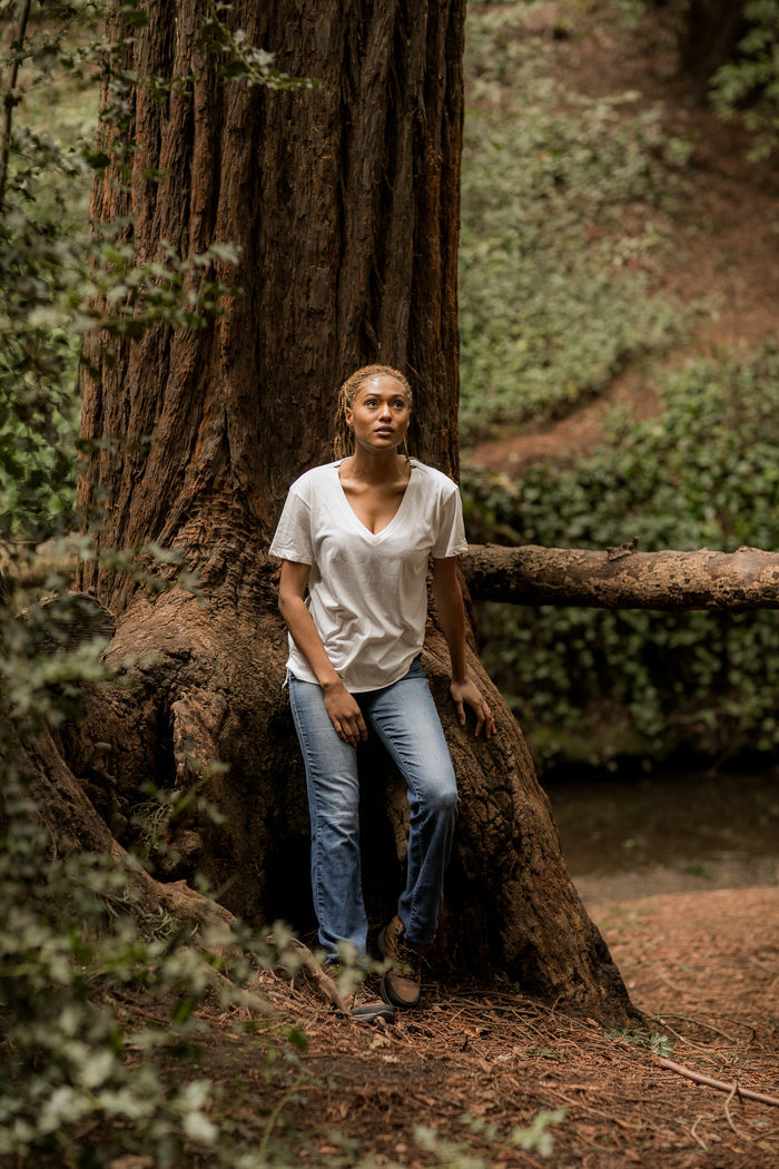 A person in a Pladra Women's V-Neck Tri-Blend Tee—Marshmallow White Heather stands by a tree in the forest.