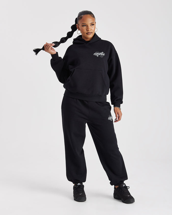 A model wearing a black hooded sweatshirt and matching sweatpants set with small white embroidered logos, posing against a light gray studio background.