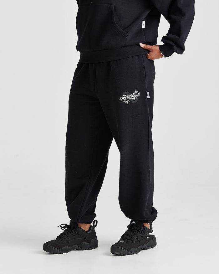 Black sweatpants with a white graphic logo on the left thigh, worn by a model with black sneakers against a light gray studio background.