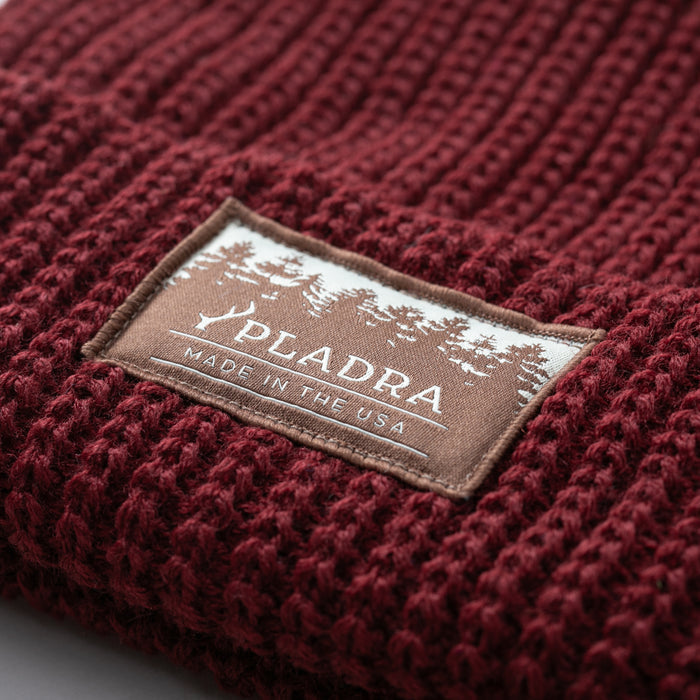 A close-up of the Woodsman Heavyweight Cuff Beanie in Burgundy Red, featuring a rectangular PLADRA MADE IN THE USA patch with tree silhouettes.