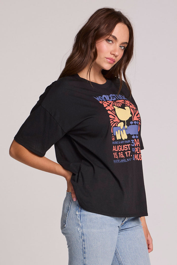 Side view of a woman wearing the Chaser  Woodstock Bird on Guitar Tee on a neutral background with hand on hip