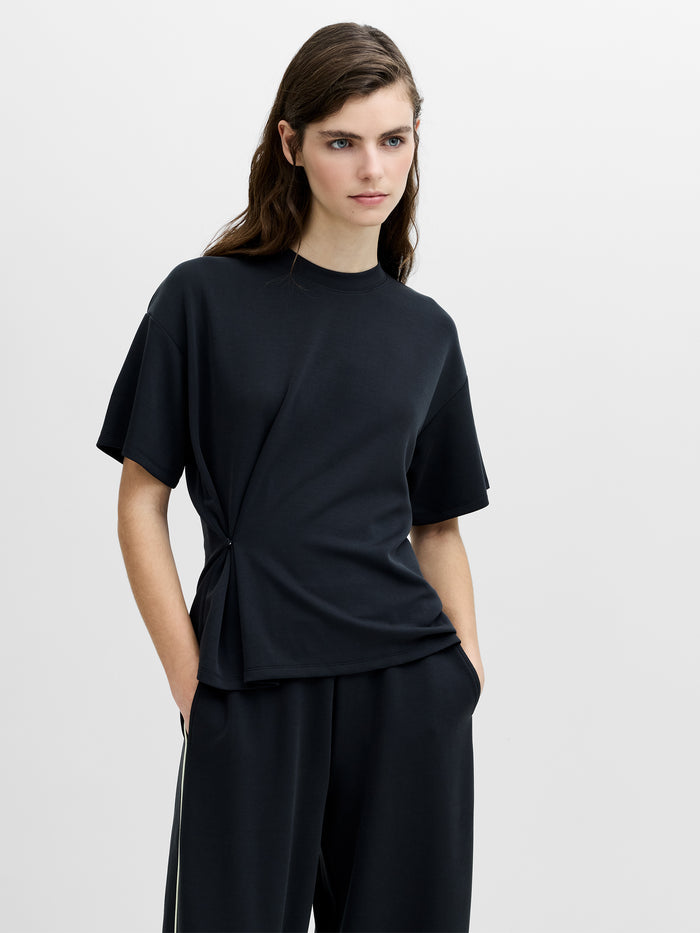 A woman wearing a dark grey relaxed fit T-shirt with a cinched waist, dropped shoulders, a crew neck and short sleeves.